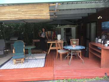 Um pátio externo aconchegante apresenta um deck de madeira, várias cadeiras, mesas, um guarda-sol de bambu, utensílios de cozinha, um tapete, plantas e uma mulher sorridente ao fundo.