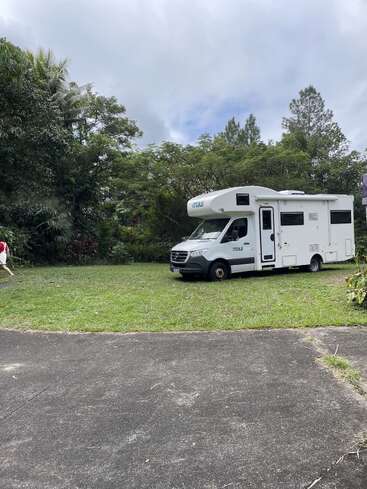 Um motorhome branco está estacionado em uma área gramada cercada por árvores verdes exuberantes, com uma pessoa de vermelho visível no lado esquerdo da imagem.