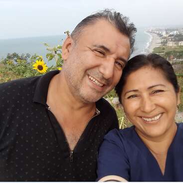 L'image représente un homme et une femme posant pour un selfie devant une vue panoramique de la côte, avec un tournesol et des bâtiments visibles à l'arrière-plan.