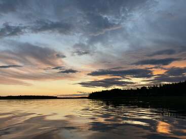 A imagem mostra um pôr do sol tranquilo sobre um lago calmo, com nuvens espetaculares pintadas em tons de laranja e azul, refletindo lindamente nas águas ondulantes.