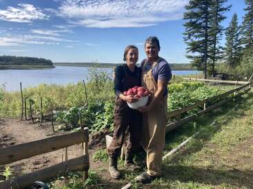 Um casal está em um jardim exuberante perto de um lago, vestindo macacões e sorrindo, segurando com orgulho uma tigela cheia de batatas vermelhas recém-colhidas.