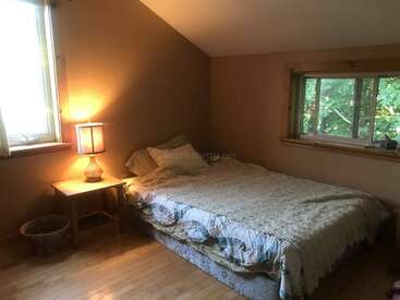 A cozy bedroom with wooden floors, a double bed with a patchwork quilt, two windows, bedside table, warm lamp, and a small wicker basket. Calm ambiance.