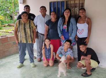 A family group of eight adults and three children pose together outside a house, smiling warmly, with a small white dog in the front, and greenery behind.