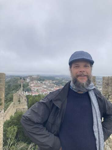 Un homme barbu, coiffé d'un chapeau et d'une écharpe, sourit sur un mur de pierre. Une vieille ville et un ciel nuageux sont visibles à l'arrière-plan.
