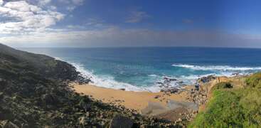 Une scène côtière à couper le souffle comprenant une plage de sable doré, des rochers escarpés, une végétation d'un vert éclatant et des vagues de l'océan d'un bleu profond sous un ciel partiellement nuageux, mariant à la perfection la beauté de la nature.
