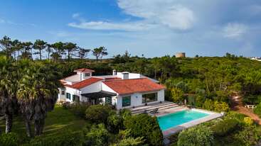 A stunning white villa with a red-tiled roof sits amidst lush greenery. A sparkling blue pool is in front, surrounded by palm trees and serene nature.