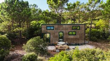 A modern tiny house with a natural, wood-colored exterior stands amidst a lush forest. Simple outdoor seating is in front, creating a peaceful, secluded retreat.