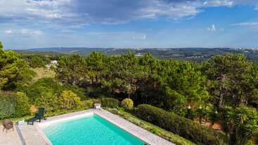 This image shows a beautiful swimming pool surrounded by lush greenery and pine trees, with scenic hills and a partly cloudy blue sky in the background. Peaceful atmosphere.