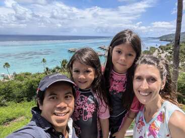Una sonriente familia de cuatro miembros posa al aire libre con el océano azul cristalino, palmeras y exuberante vegetación de fondo bajo un cielo brillante y parcialmente nublado. Ambiente vacacional.