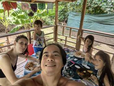 Una familia de cinco miembros disfruta de una comida en un patio cubierto rodeado de exuberante vegetación, sonriendo y compartiendo un momento feliz al aire libre en plena naturaleza.