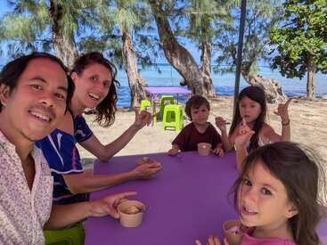 Una feliz familia de cinco miembros disfruta de un helado en una mesa violeta al aire libre junto a la playa, rodeada de árboles y agua azul claro, sonriendo juntos.