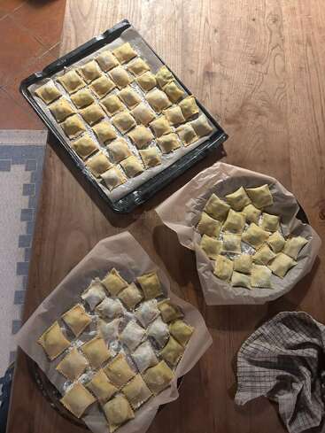 Three trays of homemade ravioli rest on parchment paper atop a wooden table, dusted with flour, ready to be cooked. A checkered cloth lies nearby.