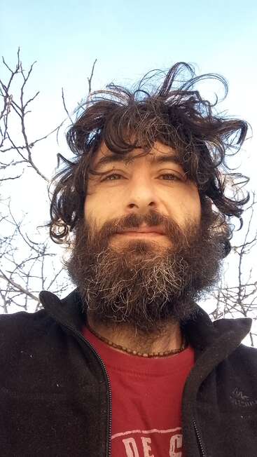A bearded man with tousled hair stands outdoors against a clear blue sky, wearing a red shirt and black jacket, surrounded by leafless tree branches.