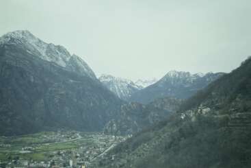 Snow-capped mountains tower over a peaceful valley dotted with houses and greenery. The village sprawls below, surrounded by rolling hills and misty, overcast skies.