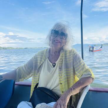An elderly woman with white hair and sunglasses smiles on a boat ride, wearing a yellow striped shirt and earrings, with calm water and blue sky behind.