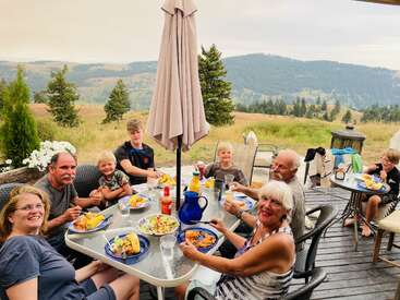 A family enjoys an outdoor meal on a patio, surrounded by nature and beautiful mountains. Everyone is smiling, sharing food, and creating joyful memories together.