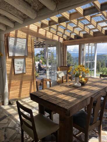 Sunlight pours into a rustic wooden dining room with a sturdy table, fresh flowers, and outdoor mountain views, where a man sits peacefully on the patio.