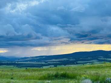 A sweeping green landscape stretches toward distant dark hills. Dramatic blue clouds fill the sky, hinting at rain, illuminated by golden sunlight peeking through. Peaceful, serene scene.