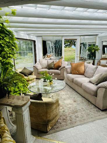 This cozy sunroom features plush couches, a round glass coffee table, soft earth-toned pillows, abundant greenery, large windows, mirrors, a patterned rug, and natural light.