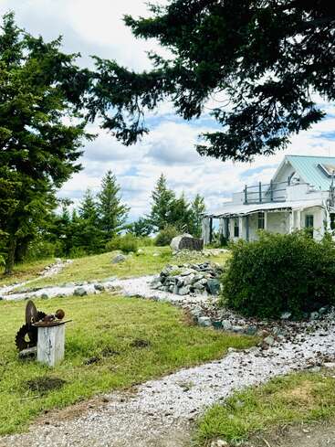 A rustic house sits among green trees and bushes, with a gravel path, old machinery, and rocks scattered across the lawn under a partly cloudy sky.