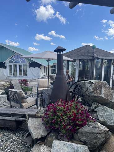 The image shows an outdoor seating area with cushioned chairs, a fireplace, large rocks, vibrant flowers, a wagon wheel, and nearby rustic buildings under a blue sky.