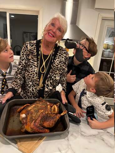 An older woman proudly presents a roasted chicken, surrounded by three children. One boy wears a fake mustache, everyone looks excited and happy in a kitchen setting.