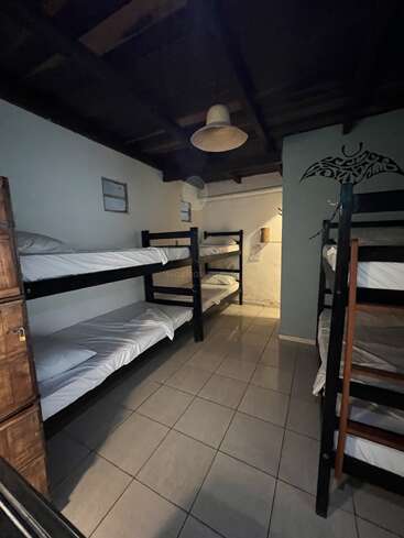 The image depicts a room with bunk beds, white sheets, and pillows, featuring a tiled floor, white walls, and a dark ceiling, with a light fixture hanging from the ceiling.