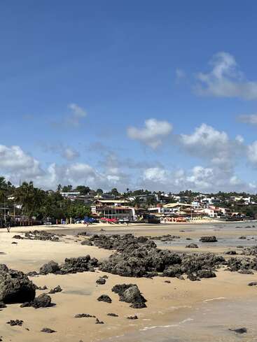 A sunny beach with scattered dark rocks, golden sand, palm trees, colorful umbrellas, and houses on a hill under a bright blue sky with fluffy clouds.