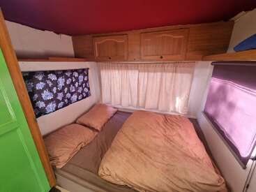Ein gemütliches, kompaktes Schlafzimmer verfügt über ein Doppelbett mit brauner Bettwäsche, zwei Kissen, Vorhänge in Blumenmuster und Lila, Hängeschränke aus Holz und cremefarbene Vorhänge vor einem Fenster.