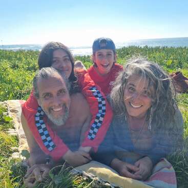 A happy family of four lies smiling together on a blanket in the grass by the seaside, enjoying a sunny, beautiful day outdoors with love and joy.