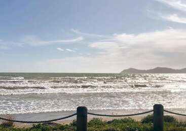 A sunny beach scene with gentle waves rolling onto the shore, distant mountains, blue sky, and a rope fence in the foreground. Calm and inviting atmosphere.