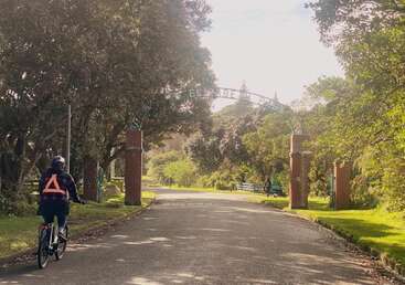 A person rides a bicycle towards a park entrance labeled "ELIZABETH PARK" on a sunny day, surrounded by trees, green grass, and peaceful paths.