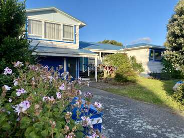 This image shows a light blue house with a blue roof, surrounded by lush greenery and colorful flowers. The driveway leads toward the welcoming entrance.