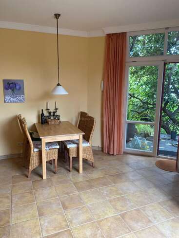 A cozy dining area with a wooden table and four wicker chairs, a hanging lamp, wall art, large windows, peach curtains, and tiled flooring.