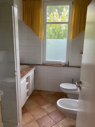 This bathroom features yellow curtains, a large window, white tiled walls, a brown countertop, a sink, toilet, bidet, and a glass shower with tiled floor.