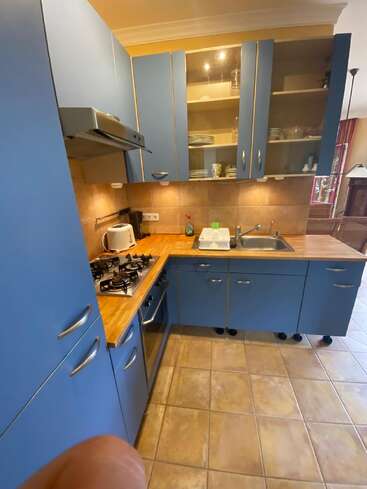 This kitchen features blue cabinets, wood countertops, a gas stove, oven, sink, toaster, and dish rack. Glass cabinets display dishes; beige tile flooring throughout.
