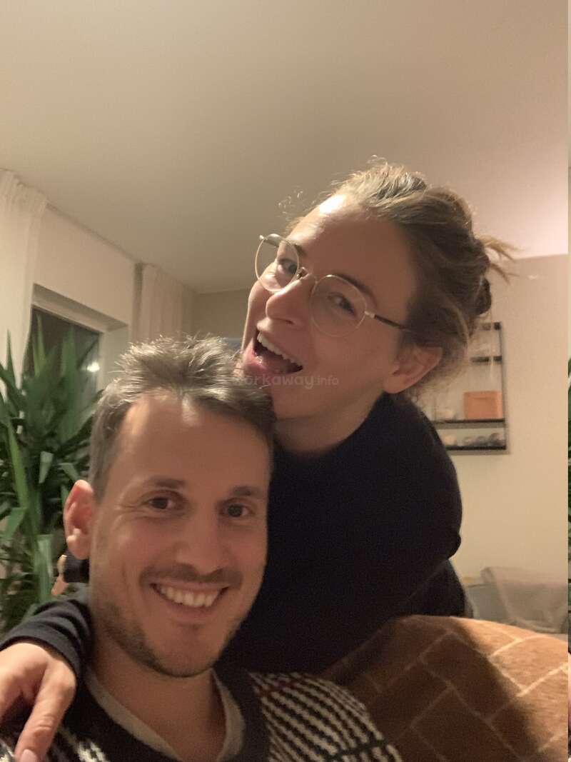 A happy couple poses indoors. The woman is playfully leaning over the man's shoulder, both smiling. Cozy room with a plant and tasteful decor in background.