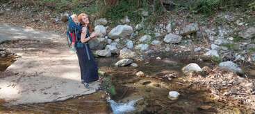 Une femme aux cheveux bouclés porte un enfant dans un sac à dos. Ils se tiennent près d'un petit ruisseau rocailleux, entourés par la nature, profitant d'une aventure en plein air.