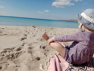 Un petit enfant portant un chapeau de soleil est assis sur une couverture sur une plage de sable, regardant la mer calme et bleue sous un ciel partiellement nuageux.