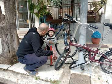 Un homme est agenouillé à côté d'un enfant souriant portant un casque. Ils gonflent un pneu de vélo sur un trottoir ensoleillé, entouré de vélos, d'arbres et de plantes.