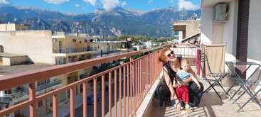 Une femme et un enfant posent joyeusement sur un balcon ensoleillé, surplombant une montagne pittoresque avec un ciel bleu et des bâtiments résidentiels. Des meubles d'extérieur les entourent.