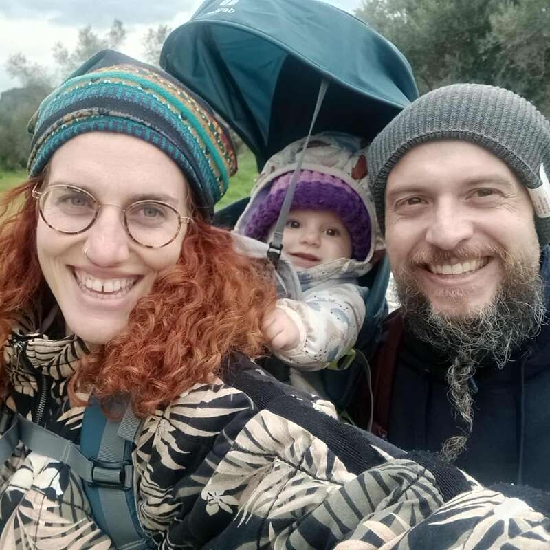Une famille souriante de trois personnes en plein air : une femme aux cheveux roux bouclés et aux lunettes, un homme barbu et un bébé dans un sac à dos.
