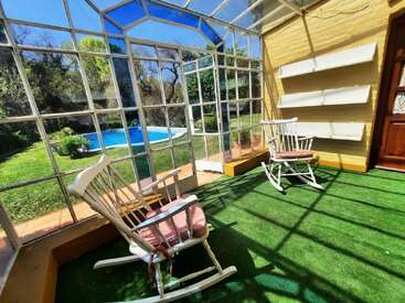 Une véranda éclairée par le soleil et dotée de parois en verre comprend deux fauteuils à bascule blancs avec des coussins, un sol en gazon artificiel, des étagères et offre une belle vue sur la piscine de l'arrière-cour.