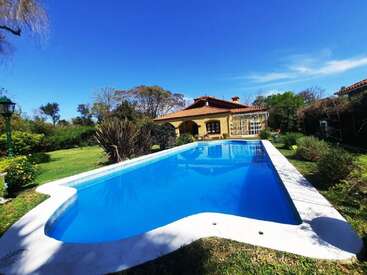 A charming house with a red tiled roof sits behind a large, inviting swimming pool, surrounded by lush greenery, trees, and a bright blue sky.