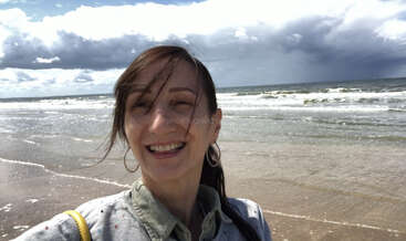 Una mujer sonriente se hace un selfie en una playa de arena con olas al fondo. El cielo nublado crea un fondo dramático y contrastado. Ella parece feliz y relajada.