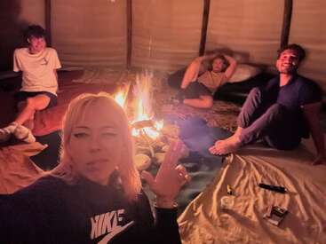 Quatro amigos relaxam em torno de uma aconchegante fogueira interna. Eles estão sentados em esteiras e cobertores, sorrindo e curtindo a companhia uns dos outros, criando uma atmosfera calorosa e descontraída.