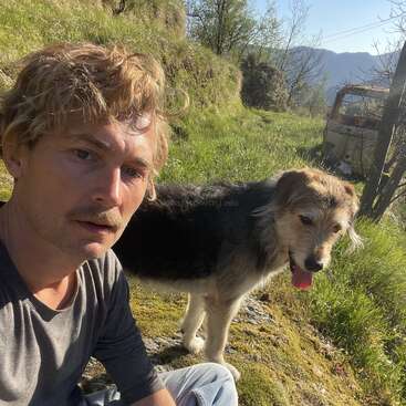 A man with blond hair and a mustache sits on grass beside a friendly dog. They are outdoors on a sunny day with green hills and trees.