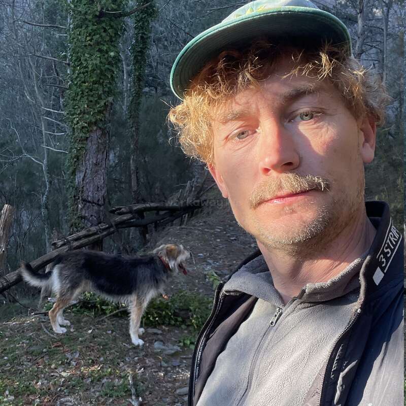 Um cara com cabelo loiro cacheado e boné tira uma selfie ao ar livre numa área florestal, enquanto um cachorro preto e branco fica atrás dele.