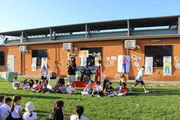 Kinder sitzen auf dem Rasen, einige stehen, vor einem orangefarbenen Gebäude, das mit bunten Zeichnungen verziert ist. Ein Erwachsener spielt Gitarre. Es scheint sich um eine Veranstaltung im Freien zu handeln.