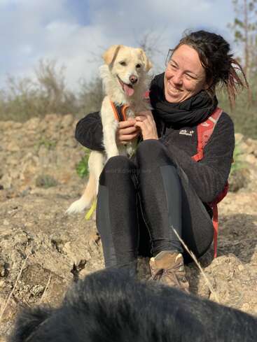 A smiling woman sits on rocky ground outdoors, holding a happy, light-colored dog in her lap. Both look content. Another animal partially visible in foreground.